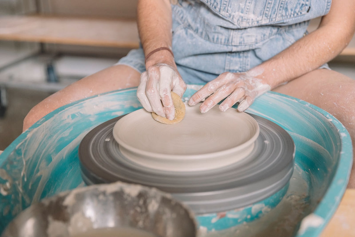 A close-up of a person's hands and torso as they shape a beige clay plate on a pottery wheel, wearing a light blue denim apron at the pottery studio sunshine coast nambour 