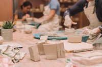 A pottery workshop scene with several people wearing aprons shaping clay bowls and blocks on a wooden table, with unfinished clay pieces in the foreground sunshine coast pottery studio nambour beginner course 