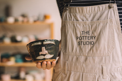 Student in The Pottery Studio apron holding a finished hand built pottery face pot, with pottery pieces in the background, Nambour Sunshine Coast